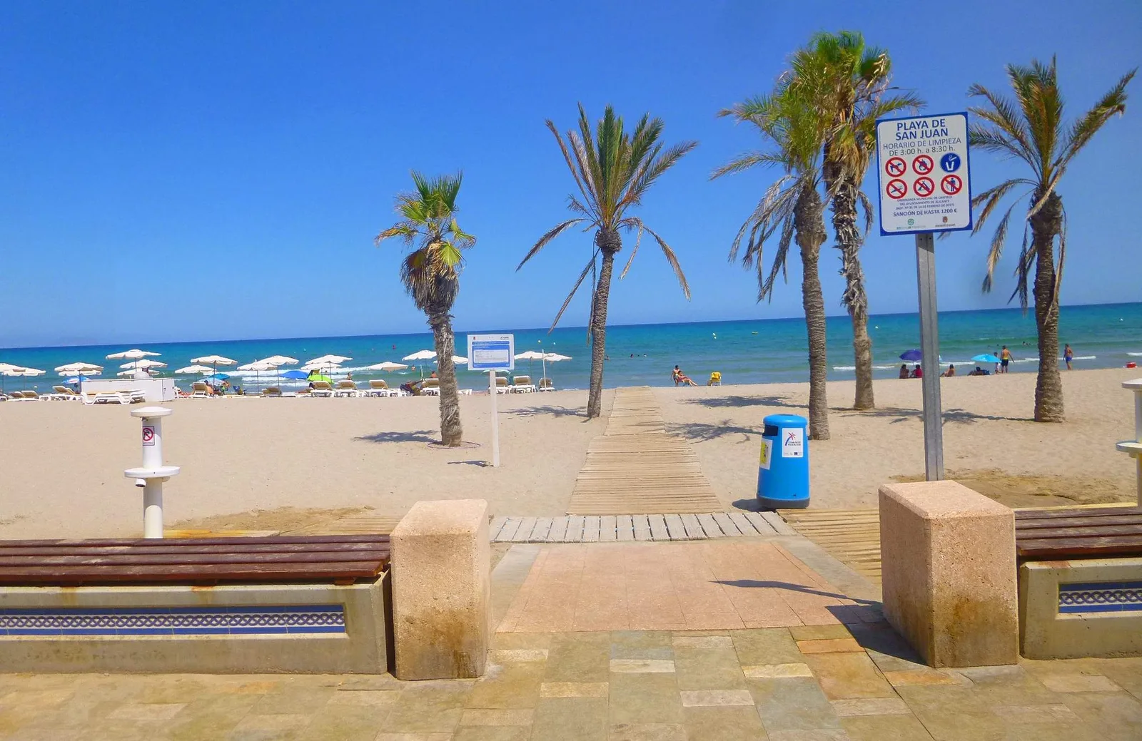 Cartel de la Playa de San Juan entre palmeras, con la pasarela de madera hacia el mar